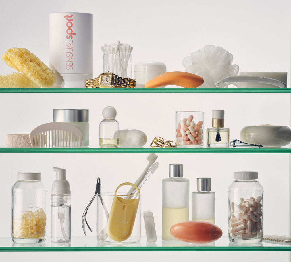 A neatly organized glass shelf displaying personal care items, including skincare products, grooming tools, supplements, a watch, and a loofah in neutral tones.