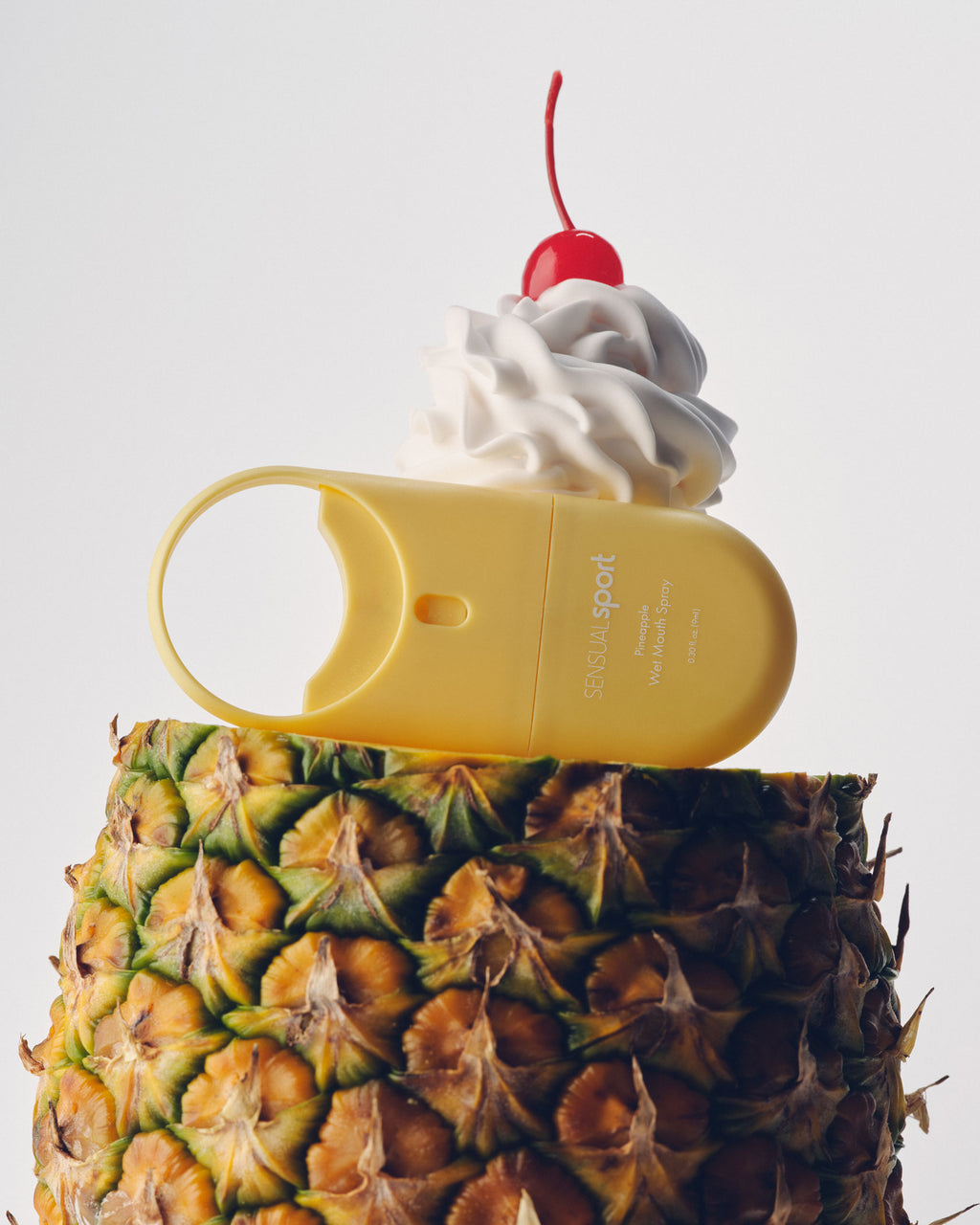 A yellow "SENSUAL sport" device rests on a pineapple, topped with whipped cream and a cherry.