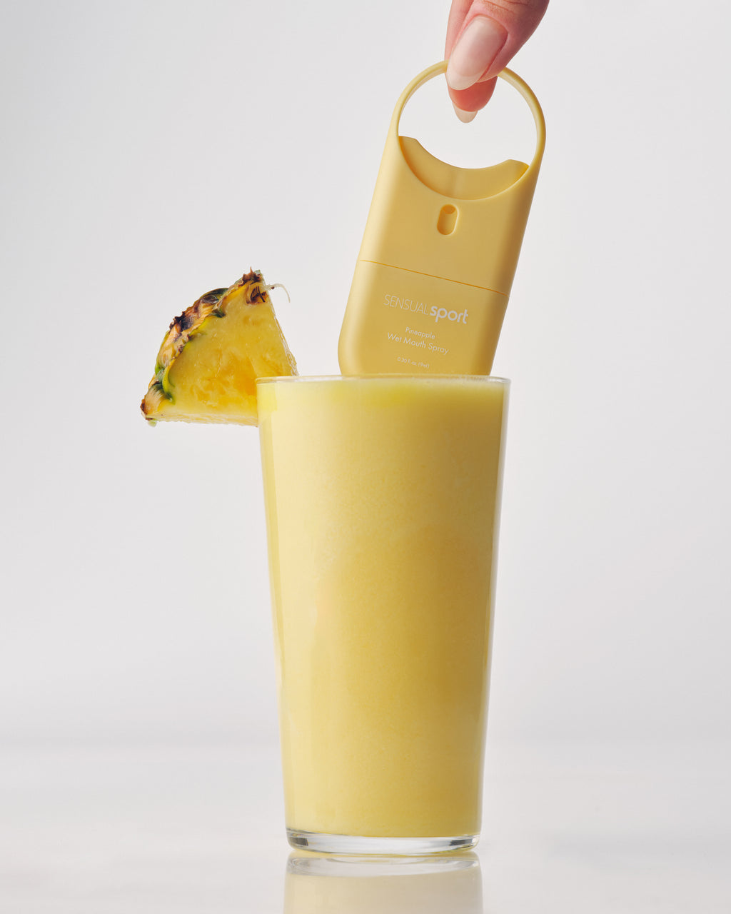 A yellow "SENSUAL sport" wet mouth spray is held above a pineapple smoothie in a tall glass, garnished with a fresh pineapple slice.