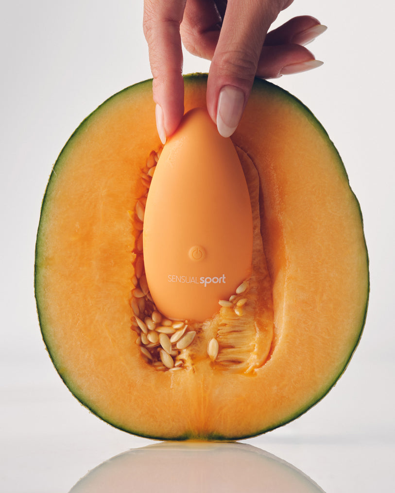 A hand pressing an orange Sensual Sport device into the center of a halved cantaloupe, blending playful and sensual imagery.